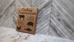A square laser-engraved oak wood magnet showing a chart of internal meat temperatures for beef, pork, and poultry, resting on a stainless steel refrigerator door