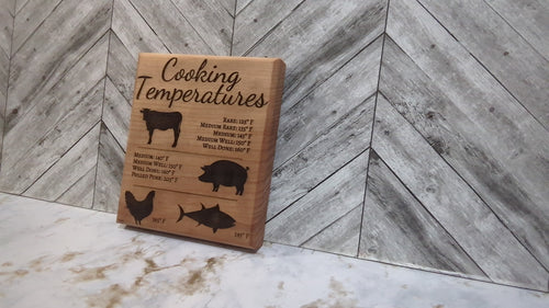 A square laser-engraved oak wood magnet showing a chart of internal meat temperatures for beef, pork, and poultry, resting on a stainless steel refrigerator door