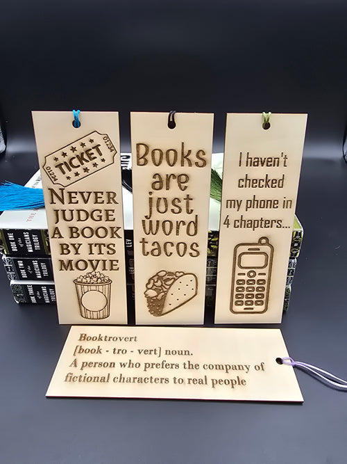 A collection of 4 laser engraved wooden bookmarks with colorful tassels that measure 6" x 2", standard bookmark size. Features laser engraved phrases that read "never judge a book by its movie" "books are just word tacos" "I haven't checked my phone in 4 chapters" "Booktrovert"