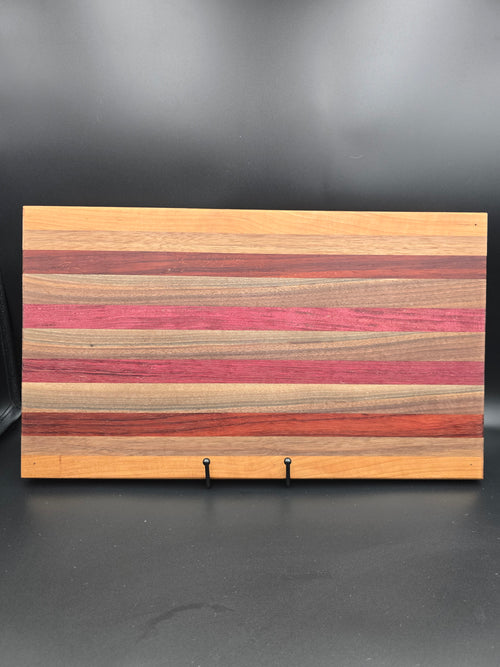 A handcrafted rectangular wooden serving board featuring a horizontal striped pattern of purpleheart, padauk, walnut, and cherry hardwoods, showcasing vibrant natural colors and a polished finish.