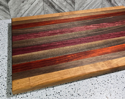 A handcrafted rectangular wooden serving board featuring a horizontal striped pattern of purpleheart, padauk, walnut, and cherry hardwoods, showcasing vibrant natural colors and a polished finish.