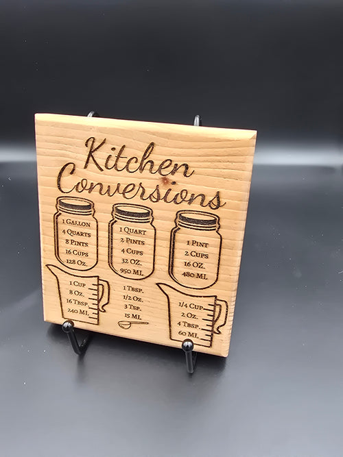 A square laser-engraved wooden kitchen conversion magnet made of a variety of hardwoods, featuring measurement charts for cups, ounces, and tablespoons, attached to a stainless steel refrigerator.
