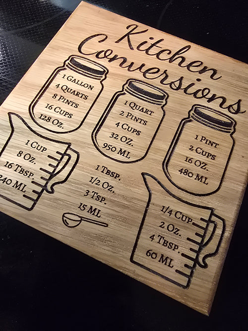 A square laser-engraved wooden kitchen conversion magnet made of a variety of hardwoods, featuring measurement charts for cups, ounces, and tablespoons, attached to a stainless steel refrigerator.