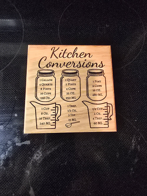 A square laser-engraved wooden kitchen conversion magnet made of a variety of hardwoods, featuring measurement charts for cups, ounces, and tablespoons, attached to a stainless steel refrigerator.